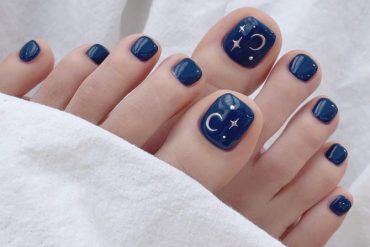 Square-shaped navy blue pedicure with crescent moon and star art on the big toes, perfect for a celestial summer nail look.