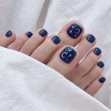 Square-shaped navy blue pedicure with crescent moon and star art on the big toes, perfect for a celestial summer nail look.