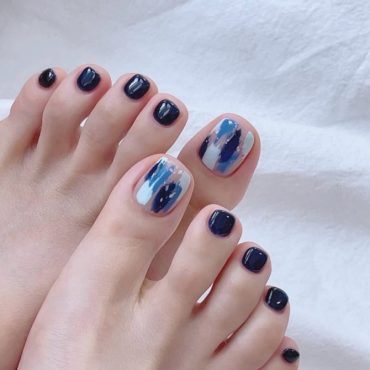 Toenails painted in deep navy blue with big toes featuring abstract brushstroke art in white, sky blue, and ink blue shades.