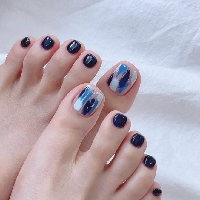 Toenails painted in deep navy blue with big toes featuring abstract brushstroke art in white, sky blue, and ink blue shades.