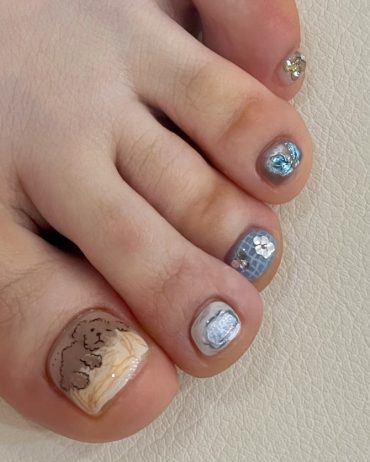 Toe nails with mixed blue shimmer textures, mosaic tiles, silver glitter, and a teddy bear design on a nude marble base