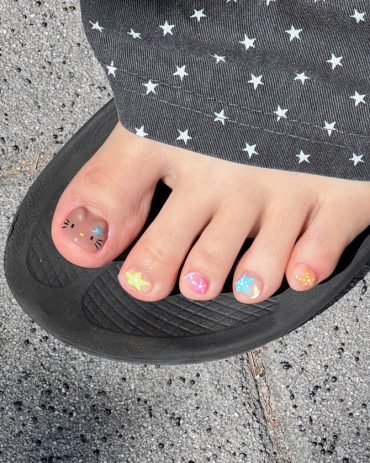 Cute rainbow-themed toe nails with a brown cat face and pastel star designs on each toe.