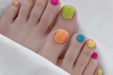 A rainbow-themed pedicure featuring glossy neon colors on each toenail including green, orange, teal, yellow, and pink.