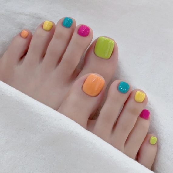 A rainbow-themed pedicure featuring glossy neon colors on each toenail including green, orange, teal, yellow, and pink.