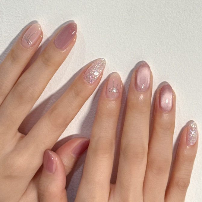Almond nails with rosy nude base, glitter accents, and a silver bow charm on one nail.