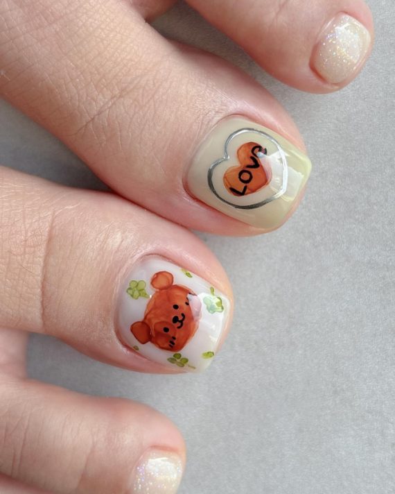 Toe nails with hand-painted teddy bear, green clovers, and double heart with “Love” text on a nude base