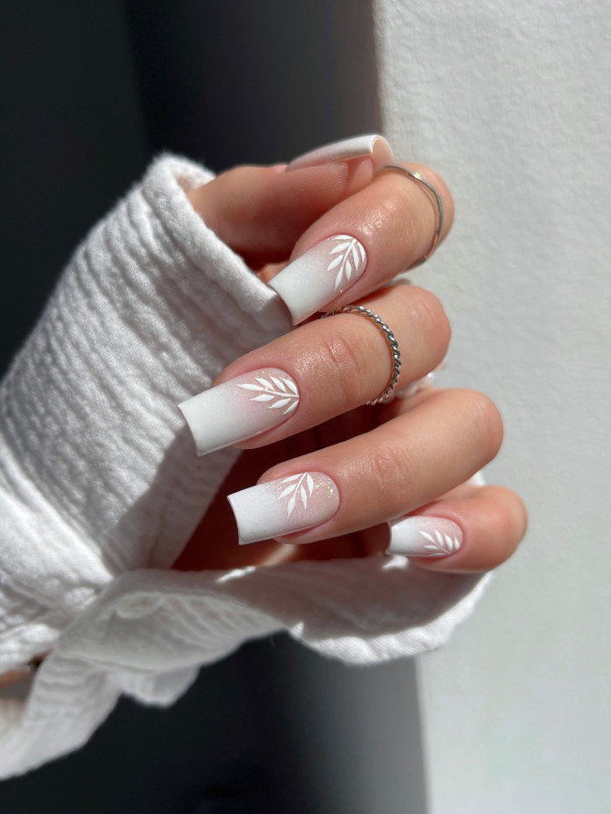 Long square matte white nails with soft ombré base and white leaf accents, worn on fair skin.