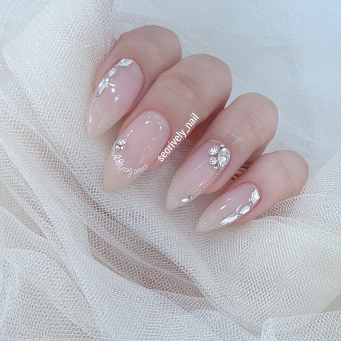 Almond-shaped nude bridal nails with clear glossy finish and sparkling crystal accents, perfect for a glamorous and timeless wedding manicure.