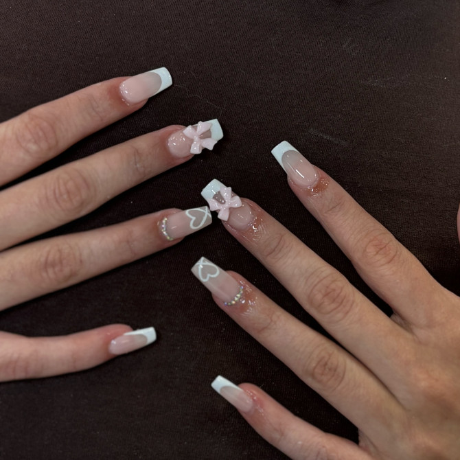 Square French tip bridal nails with glossy nude base, 3D pink bows, dainty white hearts, and subtle glitter bands—perfect for a playful yet elegant wedding style.
