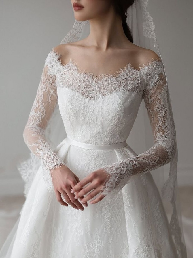 Close-up of off-shoulder lace bodice with long sleeves and slim satin belt on an A-line bridal gown. Off-shoulder floral ballgown with bishop sleeves; romantic bridal gowns mood
