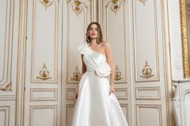 Satin one-shoulder A-line wedding gown with large rosettes at shoulder and waist in a gilded room. “Satin one-shoulder A-line inspired by international bridal gowns; large rosettes in a gilded room.