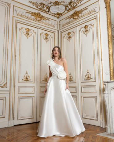 Satin one-shoulder A-line wedding gown with large rosettes at shoulder and waist in a gilded room. “Satin one-shoulder A-line inspired by international bridal gowns; large rosettes in a gilded room.