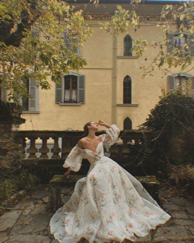 shoulder floral ballgown with bishop sleeves in a garden courtyard. Satin one-shoulder A-line inspired by international bridal gowns; large rosettes in a gilded room